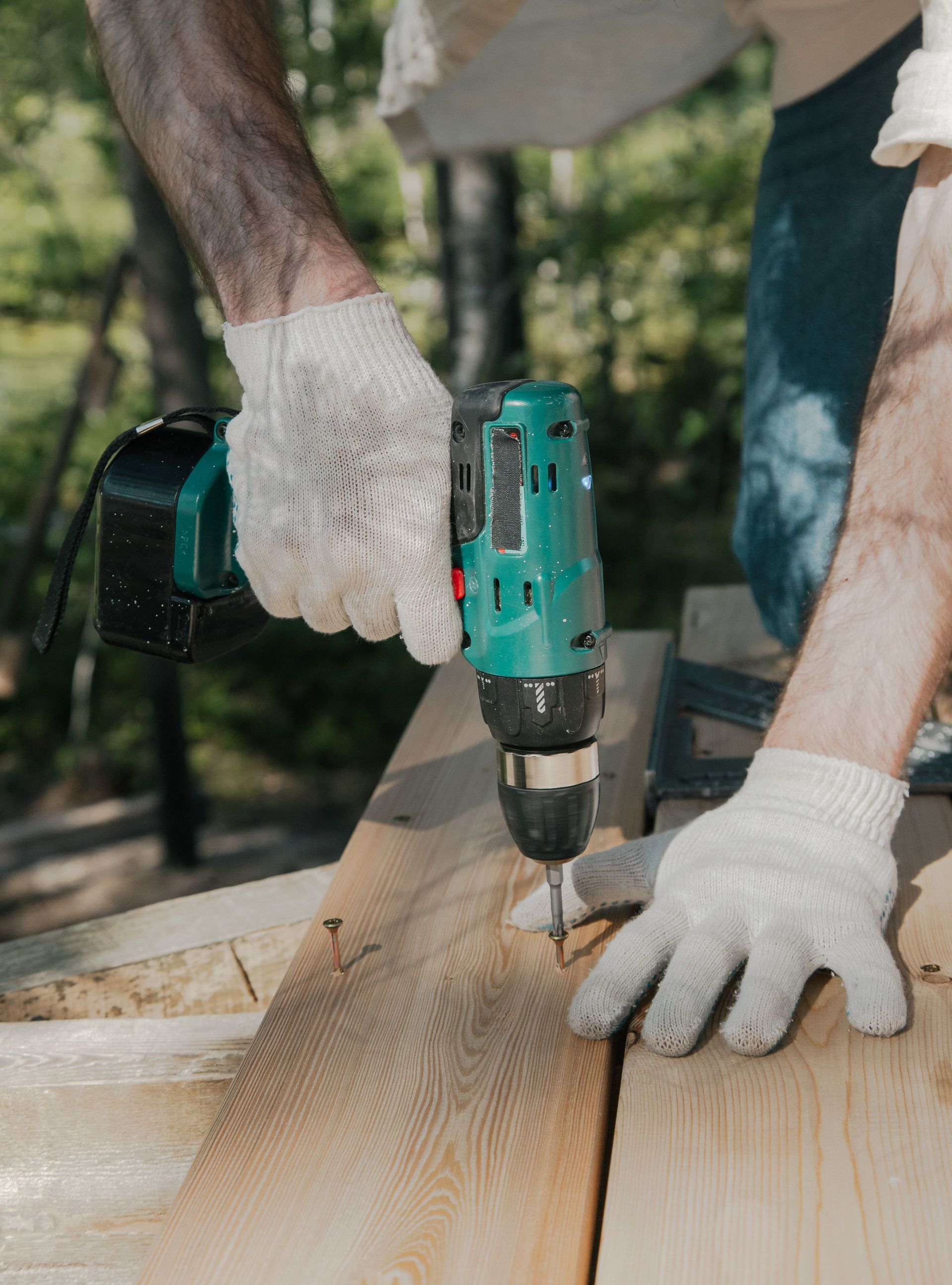 Adult using a power drill on a wooden surface, wearing white gloves, outdoors in daylight.